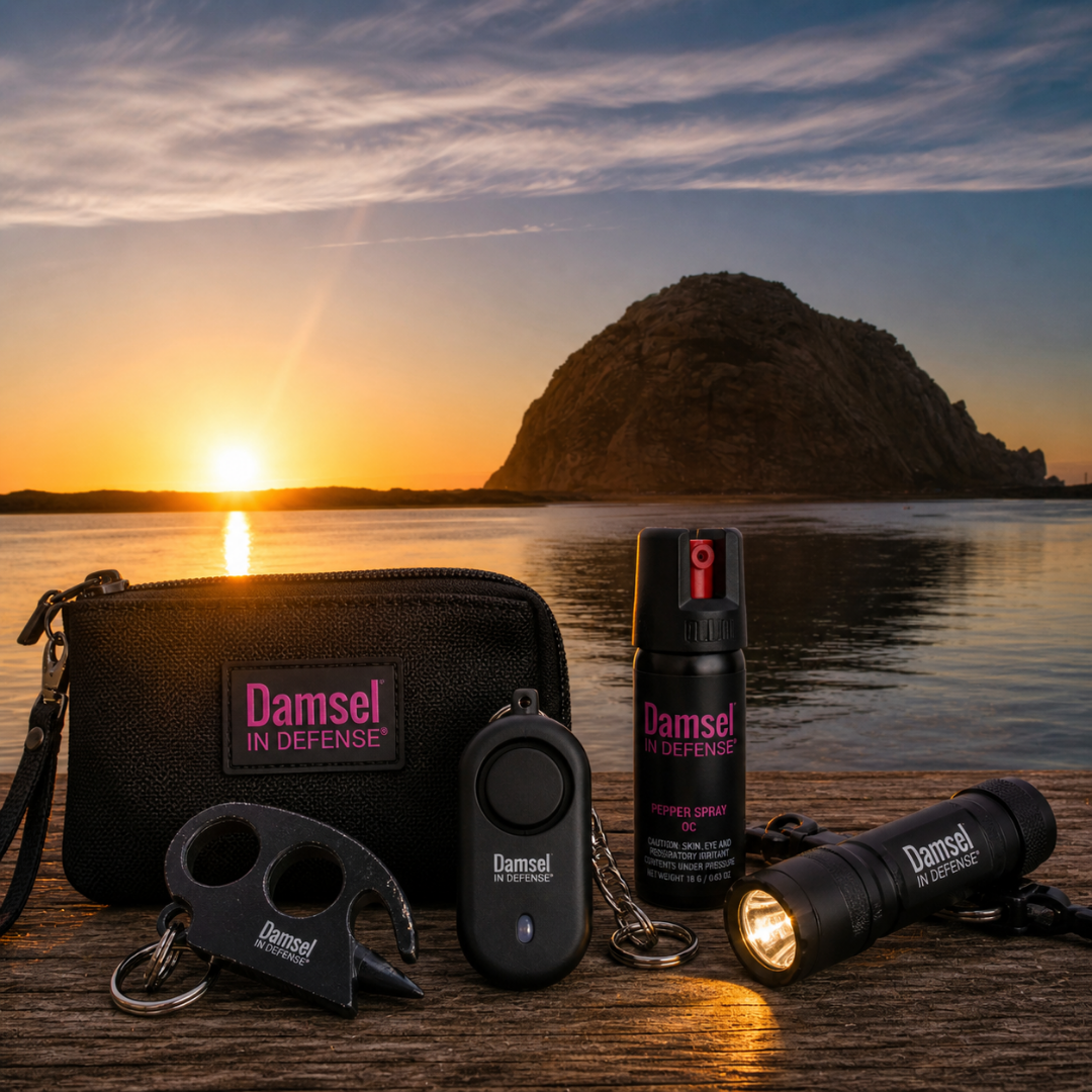 Damsel in Defense personal safety products including pepper spray, flashlight, alarm keychain, and self-defense tools displayed near Morro Rock at sunset.